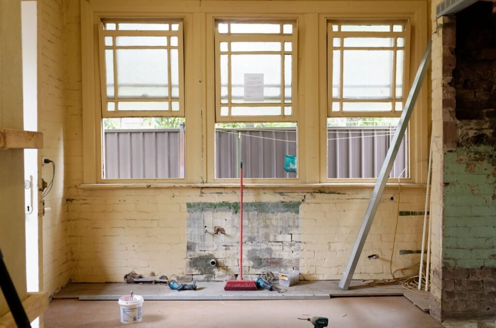 Empty room under renovation with exposed wall surfaces, tools, and three large windows.